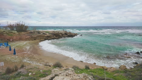 Eine-Ruhige-Strandszene-Mit-Verstreuten-Blauen-Sonnenschirmen-Und-Ohne-Menschen,-Die-Vor-Dem-Hintergrund-Des-Stürmischen-Meeres-Einen-Heiteren-Und-Verlassenen-Eindruck-Macht