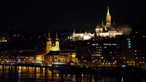 Budapest-city-center-view-with-the-illuminated-church-on-the-top-of-the-hill,-Danube-river-at-night,-gothic-architecture,-light-reflections,-panoramic-shot