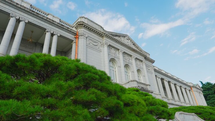 Inquadratura dal basso verso l'alto della maestosa facciata principale colonnata del Palazzo di Akasaka a Tokyo che si erge sopra una vegetazione lussureggiante e un cielo blu