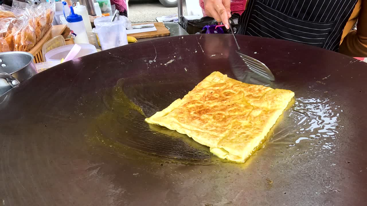 A street vendor expertly cooks fried roti on a large griddle in Kanchanaburi, Thailand. Bright lighting highlights the golden-brown texture