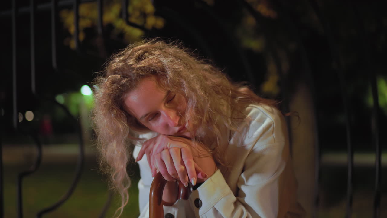 Sleepy woman with curly hair wearing white coat resting head on hands placed over umbrella handle while sitting outdoors at night under soft lights feeling tired alone