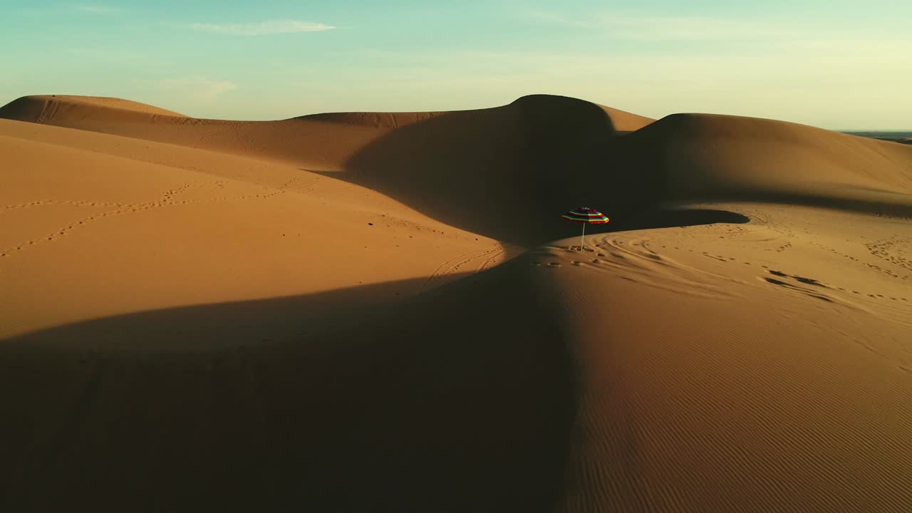 Aerial fly by of colorful beach umbrella isolated in desert sand dunes at sunset