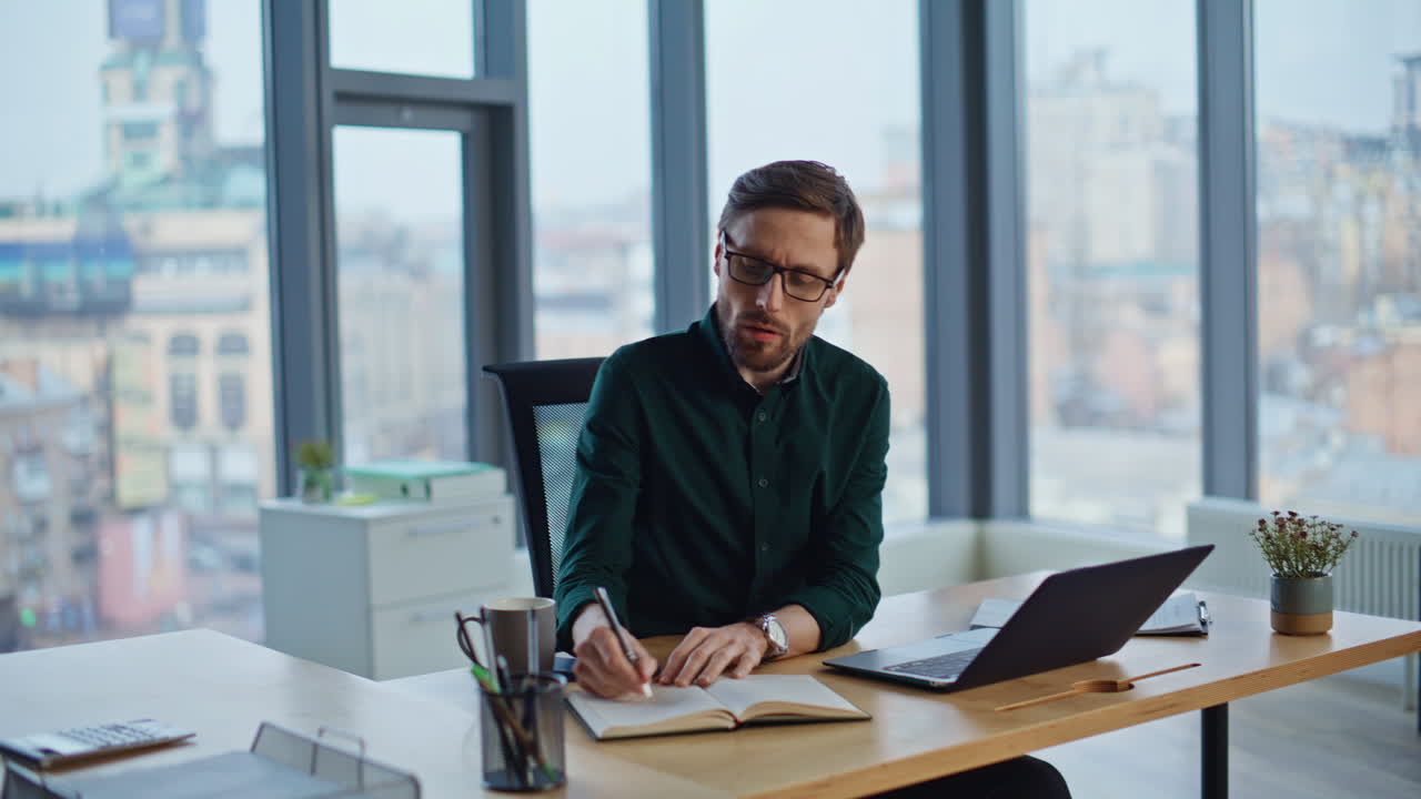 Busy employee writing notes in notebook making online research by laptop office