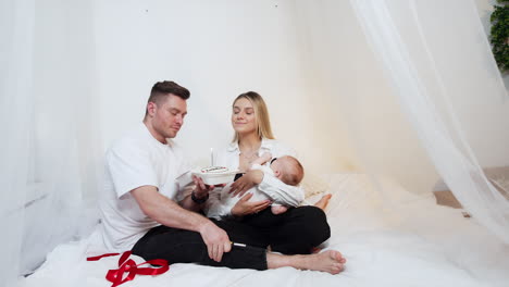lovely couple with a baby sits on the canopy bed. man holds a cake and lights the candle. parents blow the candle together making a wish.