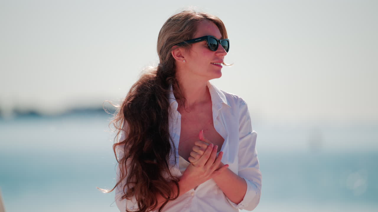 Smiling woman wearing sunglasses and a white shirt stands on the beach under the sun, with the sea glittering behind her