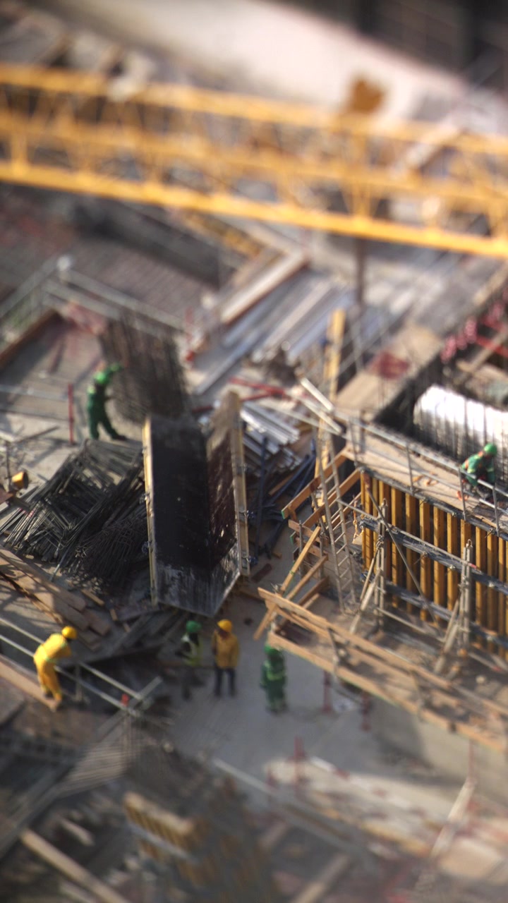 Timelapse sudut tinggi dari lokasi konstruksi yang sibuk di Dubai, menampilkan efek miniatur tilt-shift saat pekerja bergerak di sekitar cetakan beton dan sebuah derek menyapu melintasi bingkai.