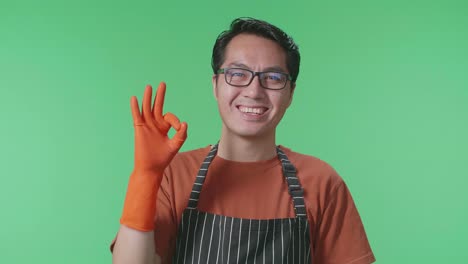 primer plano de una ama de casa masculina asiática con un delantal mostrando un gesto de ok y sonriendo mientras está de pie en el estudio de pantalla verde