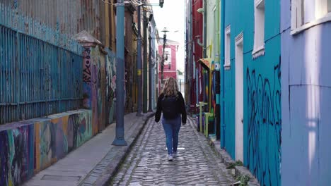 curly-haired blonde tourist girl walking with her back to the camera through a graffitied urban alley in valparaiso, chile