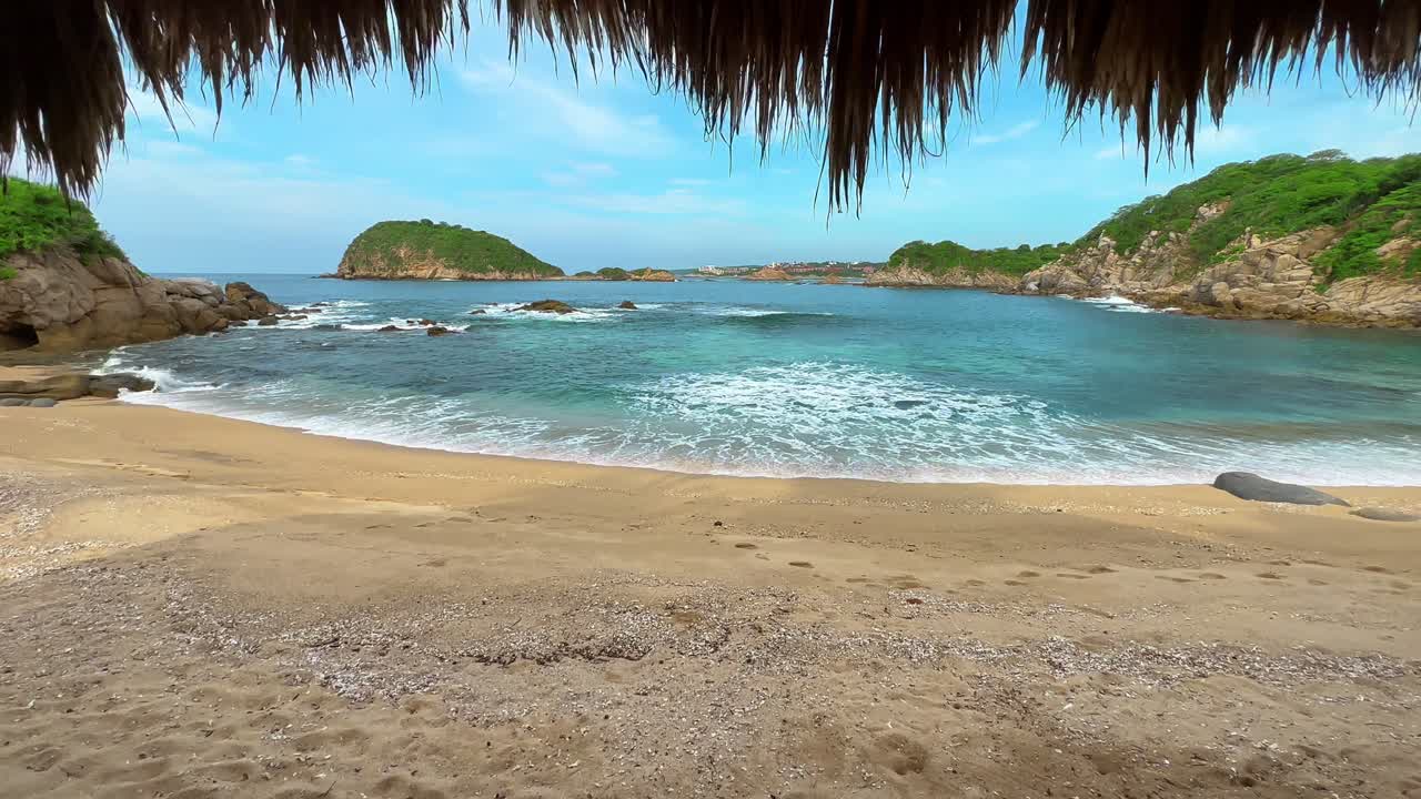 Vista aislada de la playa desde una villa con tejado de paja en Huatulco, México, con agua turquesa, olas suaves y orilla arenosa para vacaciones y escapadas tropicales.