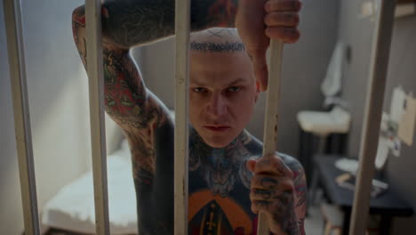 portrait of gritty tattooed inmate looking at camera through bars of prison cell