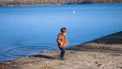 Caucasian boy throws the big stone into the river. Kid goes to find a new stone and throw it into water.