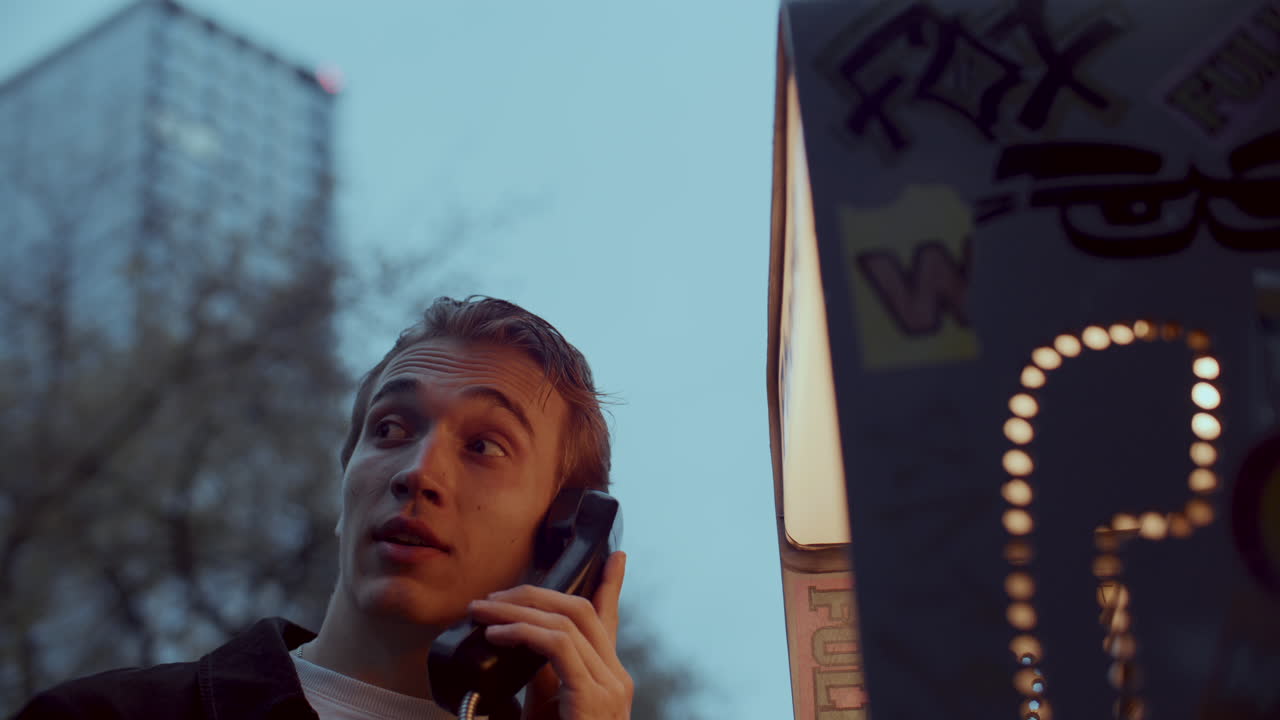 Young Guy Speaking at Public Phone Booth at Dusk in City