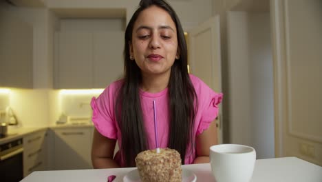 happy brunette indian girl in a pink dress blows out a candle on a small cake and claps her hands while celebrating her birthday in a modern apartment at a white table