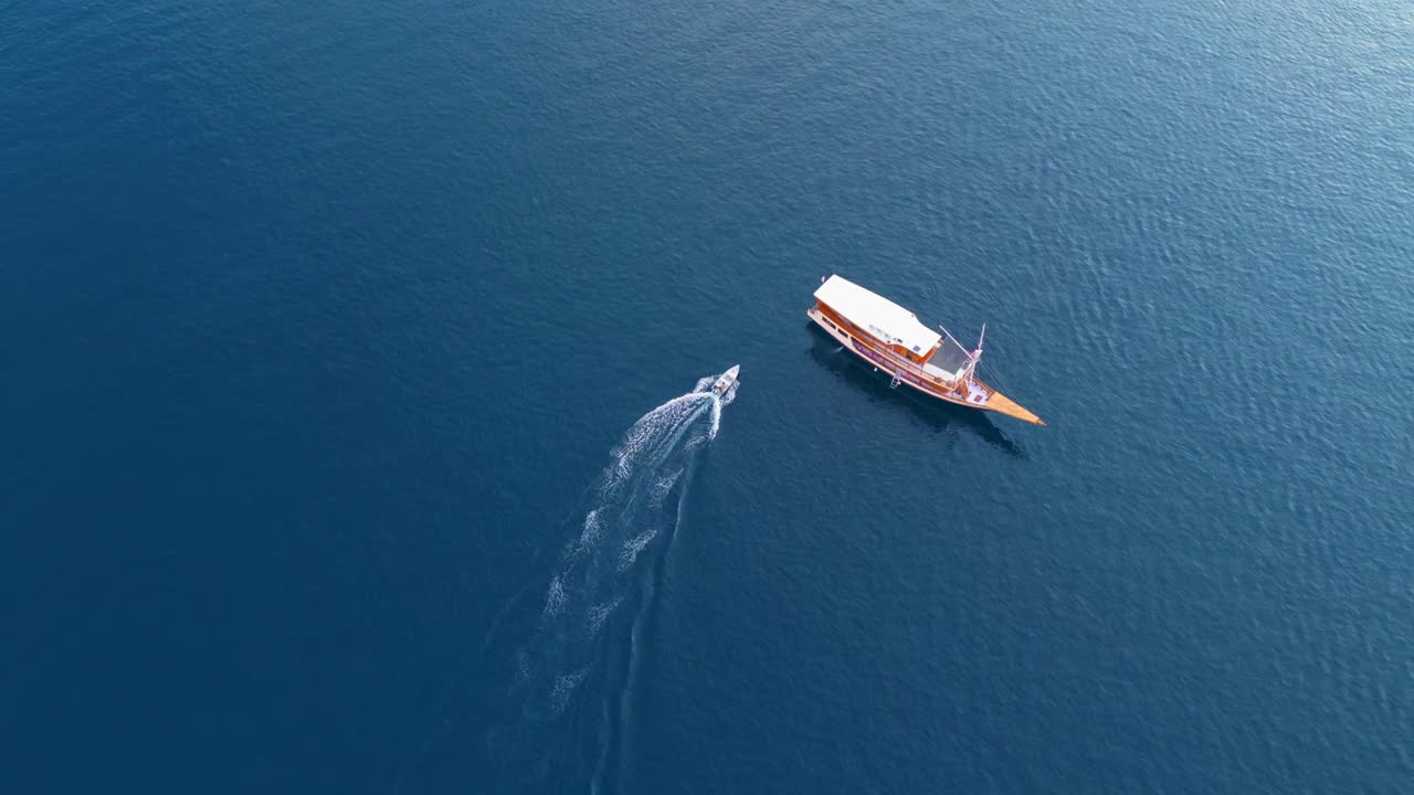 High aerial of a speedboat heading towards a large anchored boat over dark blue ocean