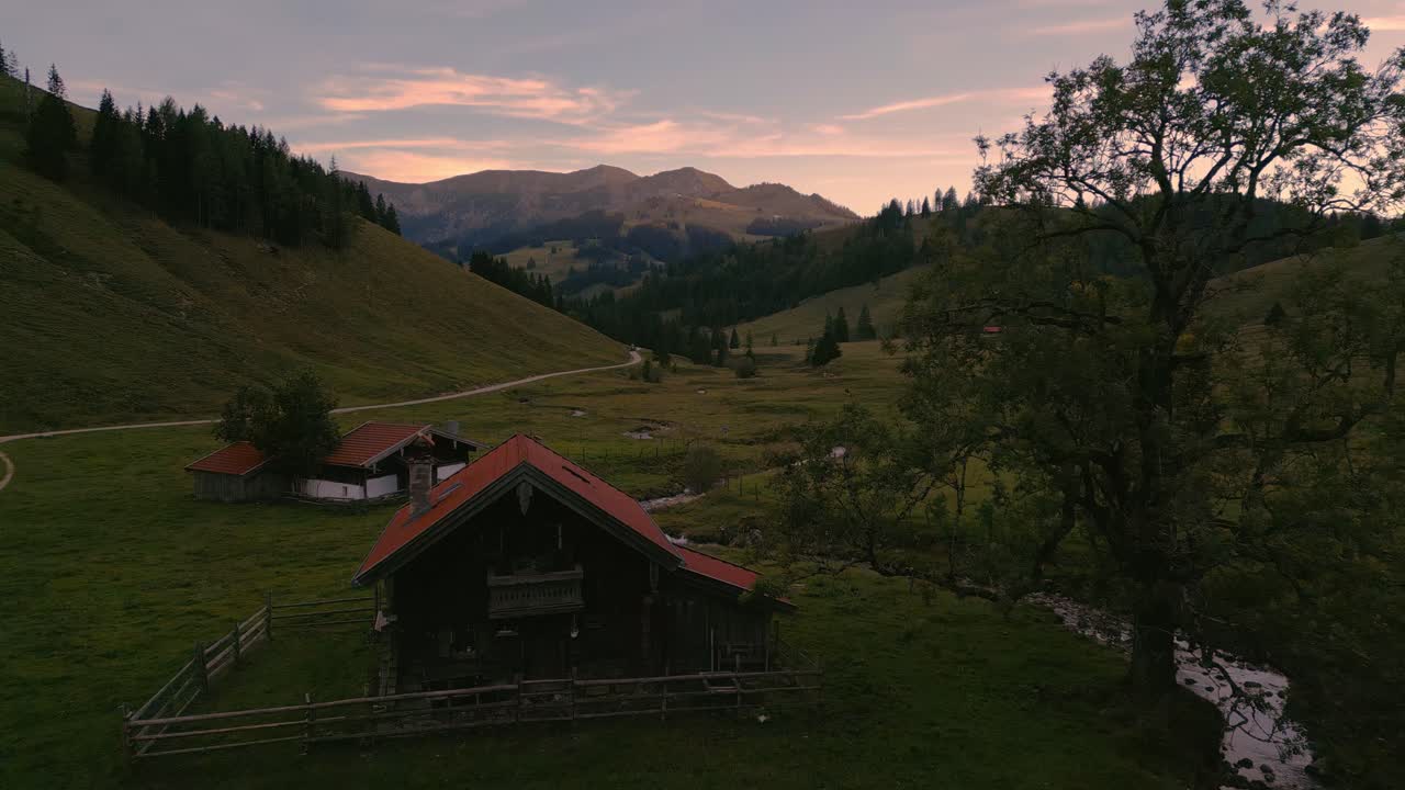 una cabaña de cabaña de madera romántica, idílica y tradicional en las montañas de los alpes bávaros al atardecer con cielo de nubes rojas, un gran árbol, prados de hierba, vacas y picos montañosos