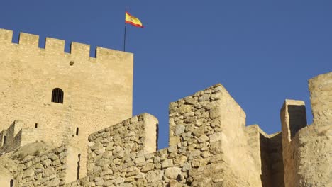 Statische-Aufnahme-Von-Unten-Auf-Die-Burg-Von-Sax-In-Alicante,-Valencia,-Mit-Der-Spanischen-Flagge,-Die-Vom-Mast-Auf-Dem-Wehrturm-Weht