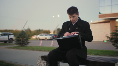 businessman seated outdoors after work, going through documents with background featuring office building, moving car, street light, and parked car in an urban setting at twilight