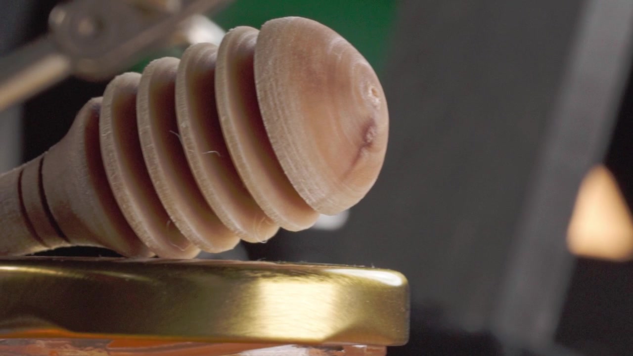 Primo piano di un mestolo di legno per miele appoggiato sul coperchio di un barattolo di vetro, che mostra le venature naturali del legno, le creste a spirale e la superficie liscia, con miele dorato visibile sotto in luce calda.