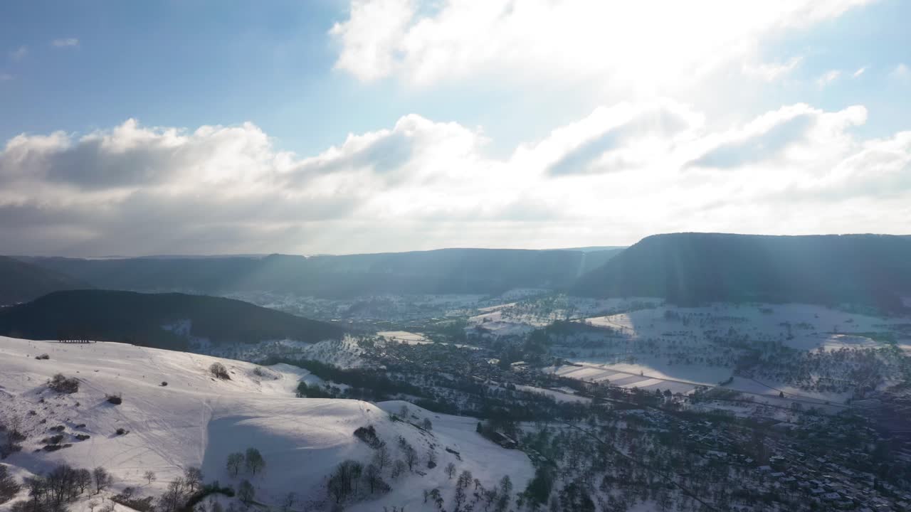 Sidelæns luftoptagelse over et vintereventyrland langs bjerge, hvor solen reflekteres i kameraet, med udsigt over et fantastisk landskab i Schwaben, Tyskland.