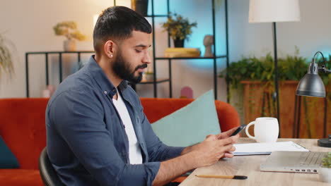 Indian-freelancer-man-using-smartphone-texting-message-while-staying-in-workplace-at-home-office