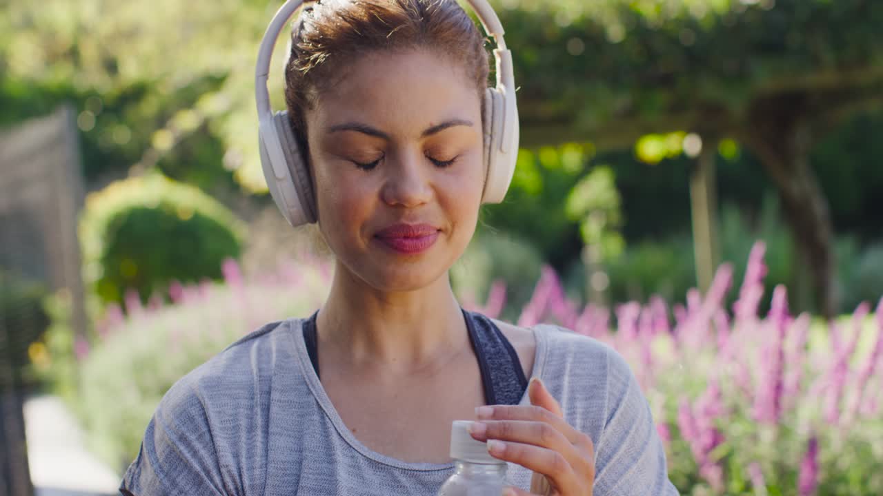 Donna afroamericana che beve da una bottiglia, controllando di chiudere bene il tappo, in abbigliamento sportivo mentre si rinfresca in giardino.