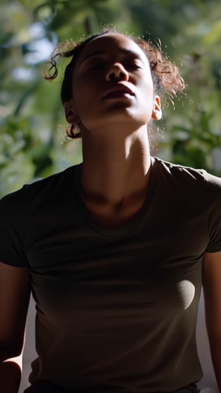 Donna si tiene la testa, occhi chiusi, in un silenzioso disagio. La luce del sole filtra attraverso le foglie verdi dietro di lei. È seduta, persa nei pensieri o nel dolore. L'immobilità suggerisce stress, stanchezza o tensione emotiva.