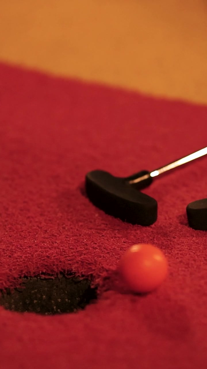 Un putter colpisce una palla arancione in una buca su un campo da minigolf rosso sotto un'illuminazione calda
