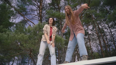 two young girl friends make fools of themselves on top of the roof of a caravan in the middle of the forest looking at the camera