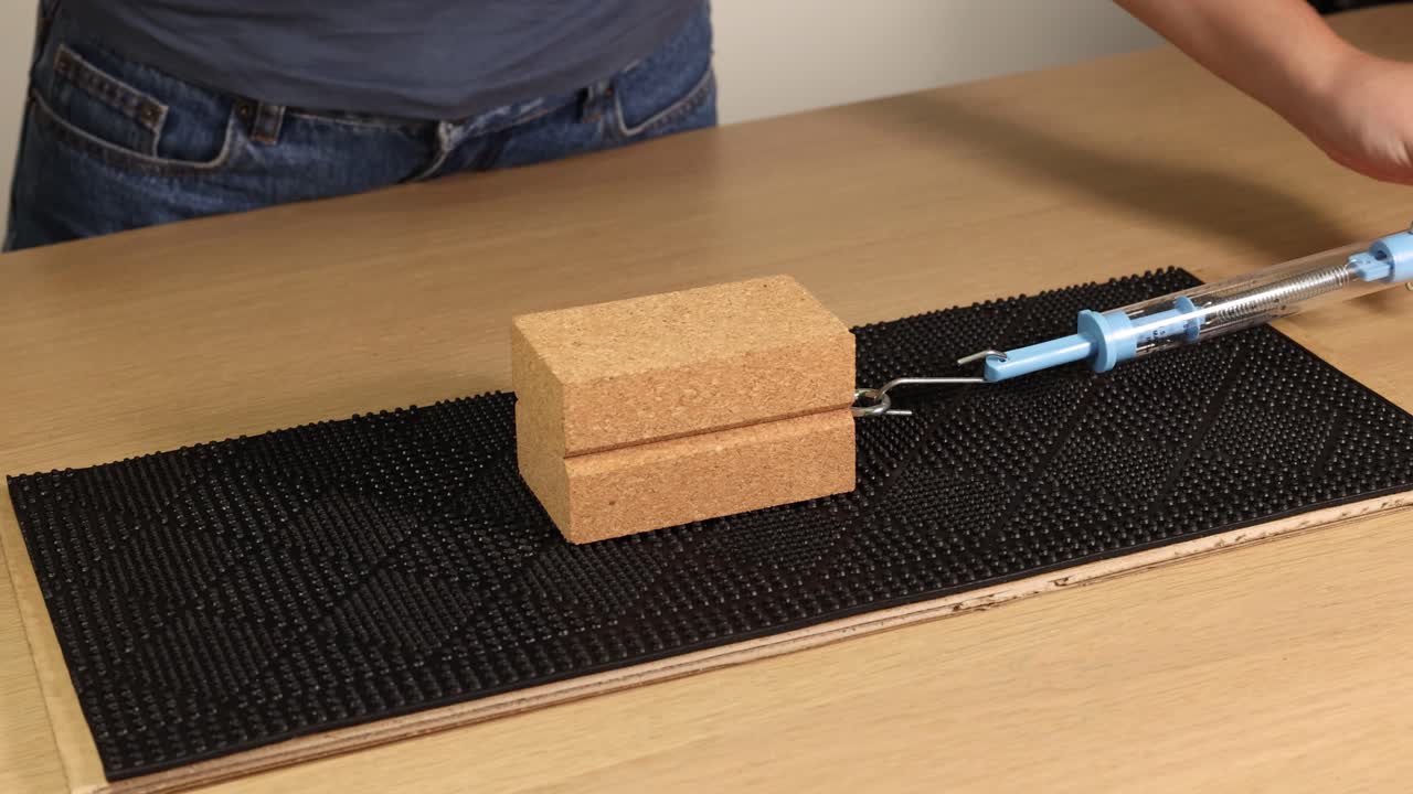 A hand pulls a wooden block using a spring scale on a textured surface, illustrating friction and force measurement