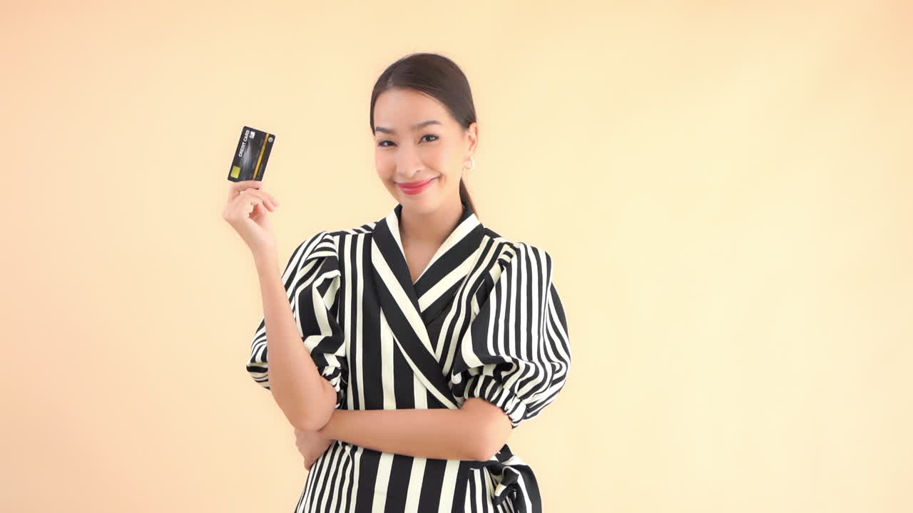 身穿黑白连衣裙的亚洲模特在纯色背景前手持信用卡或借记卡摆姿势。