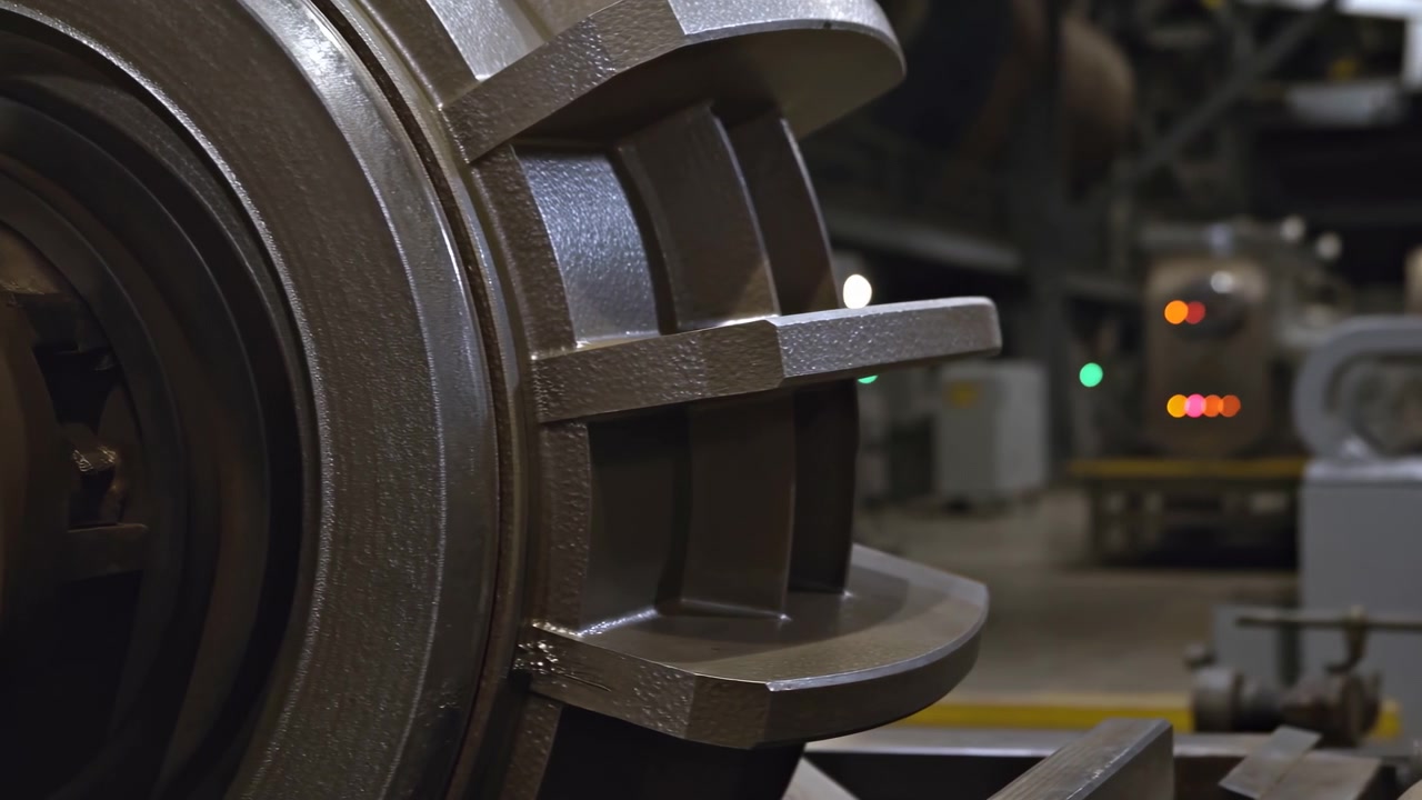 Detail velkých kovových ozubených kol a mechanických součástí v tlumeném továrním prostředí, s rozmazanými kontrolkami v pozadí naznačujícími aktivitu.