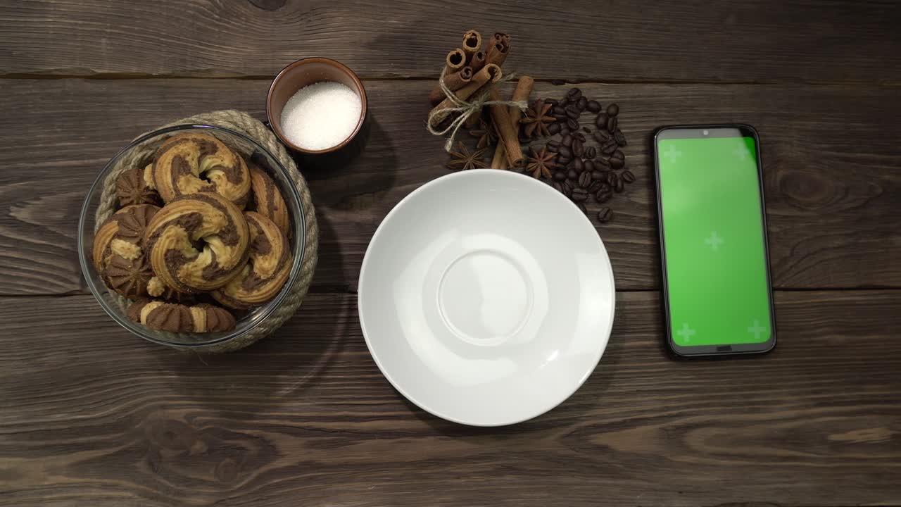 una mano femenina pone una taza de capuchino en una mesa de madera con un teléfono inteligente tendido sobre ella.