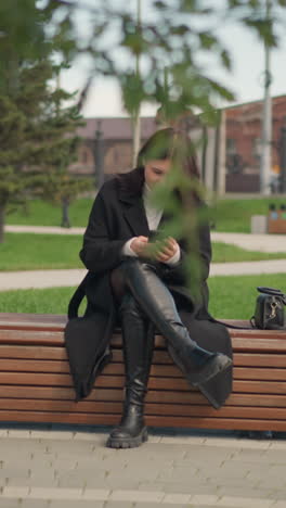 mujer sentada en un banco del parque usando un teléfono inteligente, vestida con abrigo y botas negras, rodeada de árboles verdes exuberantes y una atmósfera al aire libre relajada en un parque urbano