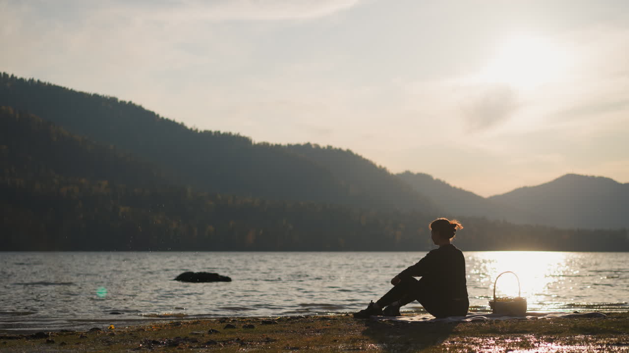 mujer lanza una roca al agua del lago al atardecer. dama silueta disfruta de un picnic en la orilla del río sola con la naturaleza rodeada por la vista nocturna de las montañas de otoño