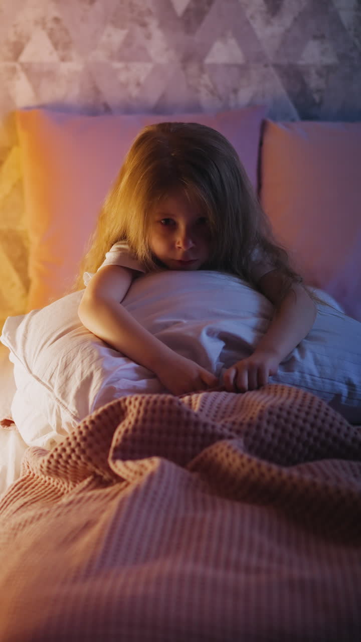 Offended blonde little girl with dimly lit face sits holding soft pillow on bed covered with pink plaid near illuminated wigwam in semidark bedroom slow motion