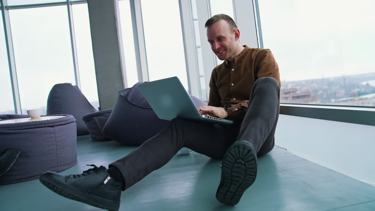 Smiling young man in modern office. Happy freelancer sitting on a floor and working on a laptop on window city view background.