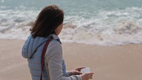 slow motion woman in warm clothes takes pictures of waves white with foam cover the wet sand of the beach on her phone, portugal