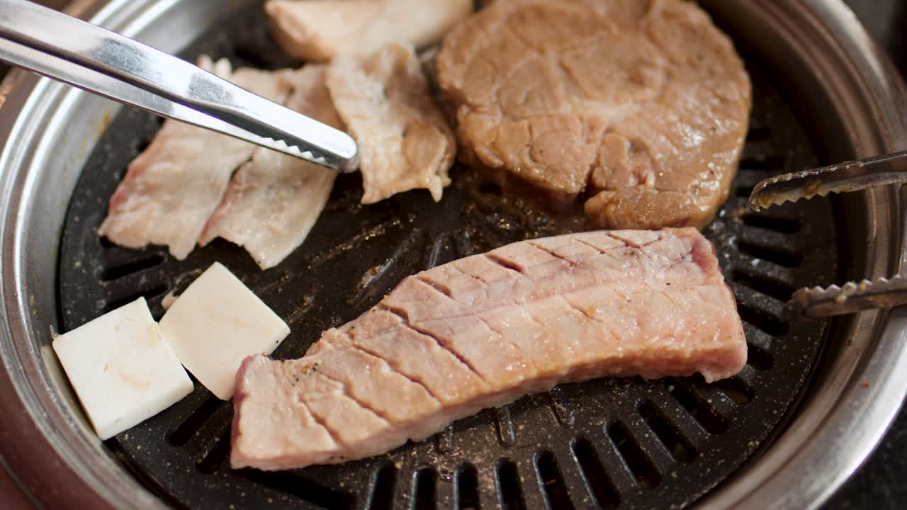 Hand uses metal tongs to arrange pork belly slices on a hot Korean barbecue grill
