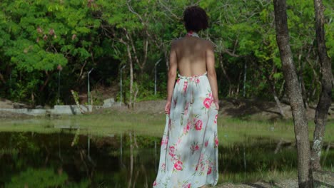 Joven-Hispana-Caminando-Hacia-Un-Lago-Con-Un-Vestido-De-Flores-En-El-Parque-En-Un-Día-Soleado