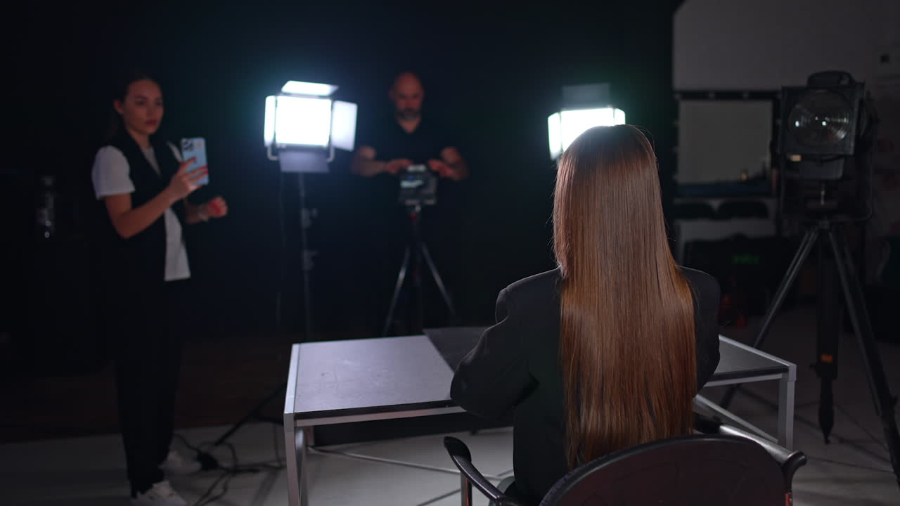 Rear view of a brunette long-haired blogger wearing a black jacket sits at desk. Woman and man at backdrop taking video on diverse devices.