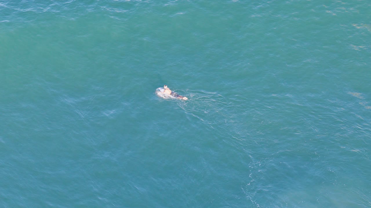 Aerial footage captures a lone surfer paddling through clear blue ocean waters near Gold Coast, Australia, under bright sunlight