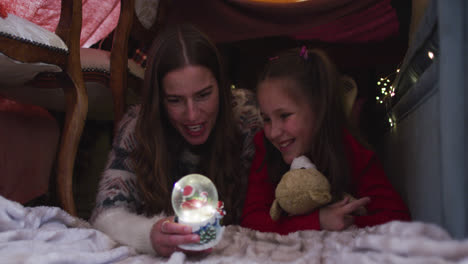 Caucasian-mother-and-daughter-holding-snow-globe-while-lying-under-blanket-fort-during-christmas-at-