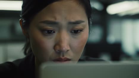 a woman sits at her desk in a dimly lit office, staring intently at her laptop screen. her facial expressions reflect concentration and concern as she navigates a critical task late at night.