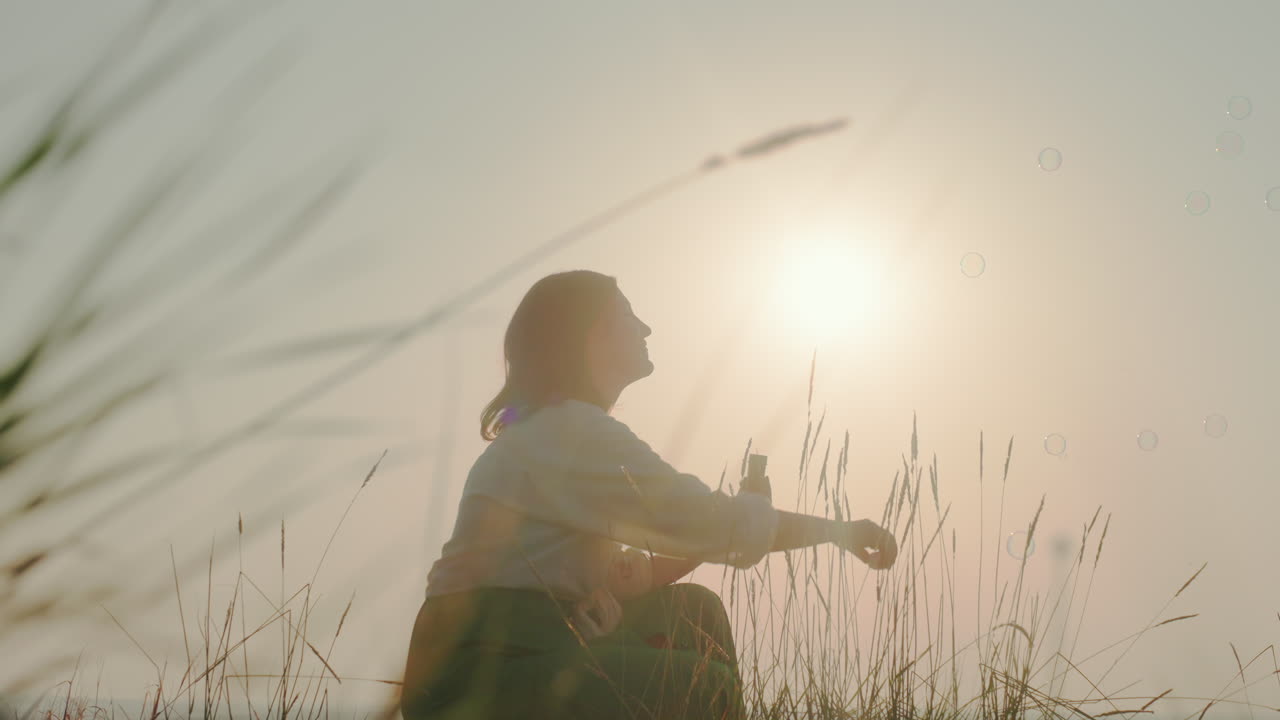 Silhouetted woman crouches in tall grass blowing bubbles toward glowing sunset sky, warm light casting long shadows across field, floating bubbles catching soft hues