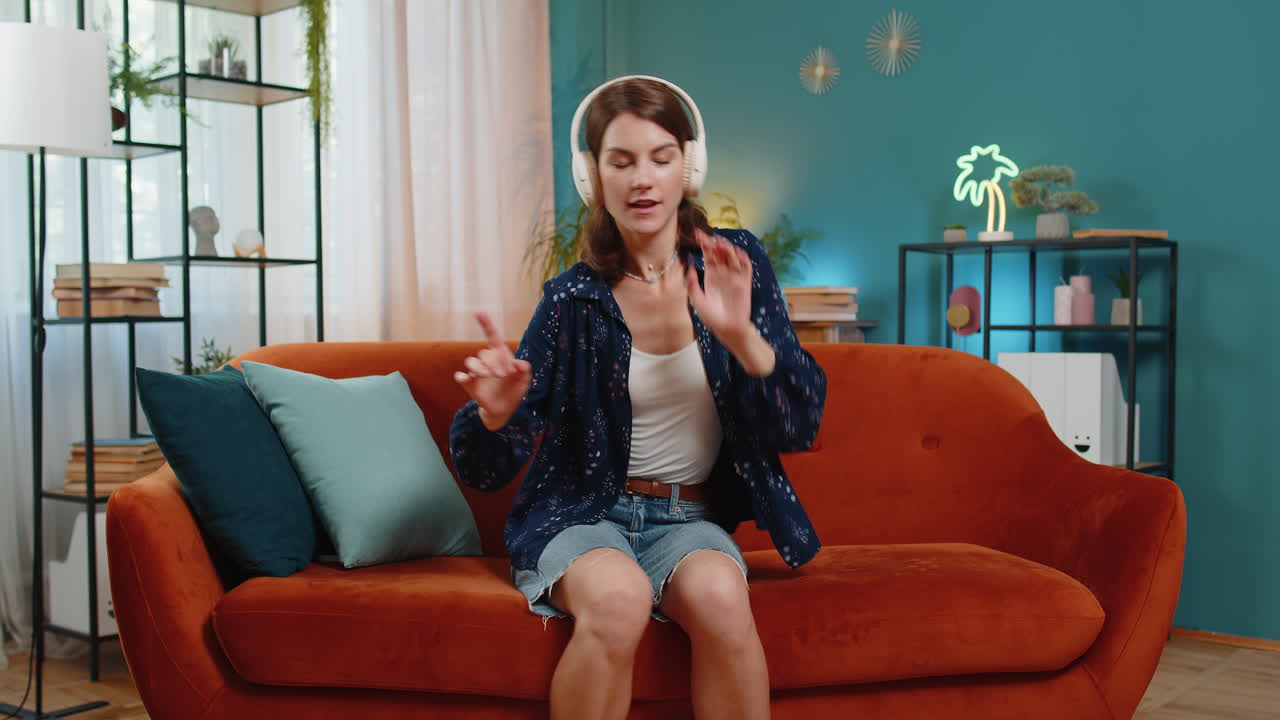 Giovane donna gioiosa che si diverte ad ascoltare musica con le cuffie wireless ballando sul divano della stanza di casa