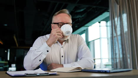 senior caucasian man in glasses sits at desk enjoying tea or coffee. a break from work in the office.