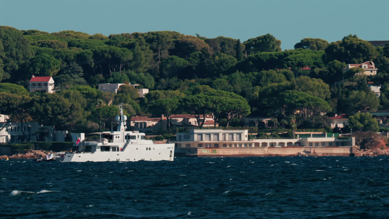 Luxury white yacht sailing near Mediterranean coastline with elegant villas surrounded by trees