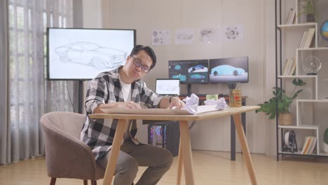 asian male crumpling paper while working on a car design sketch on table in the studio with tv and computers display 3d electric car model