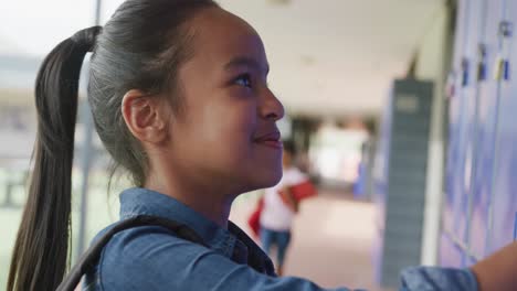 Video-portrait-of-happy-biracial-schoolgirl-using-locker,-smiling-in-school-corridor,-copy-space