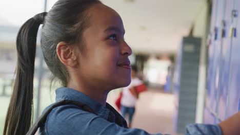 video portrait of happy biracial schoolgirl using locker, smiling in school corridor, copy space